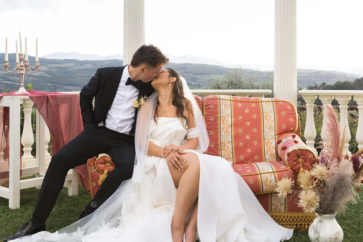 Newlyweds kiss while sitting on a sofa in front of the colonnade of the Monastero Santa Margherita