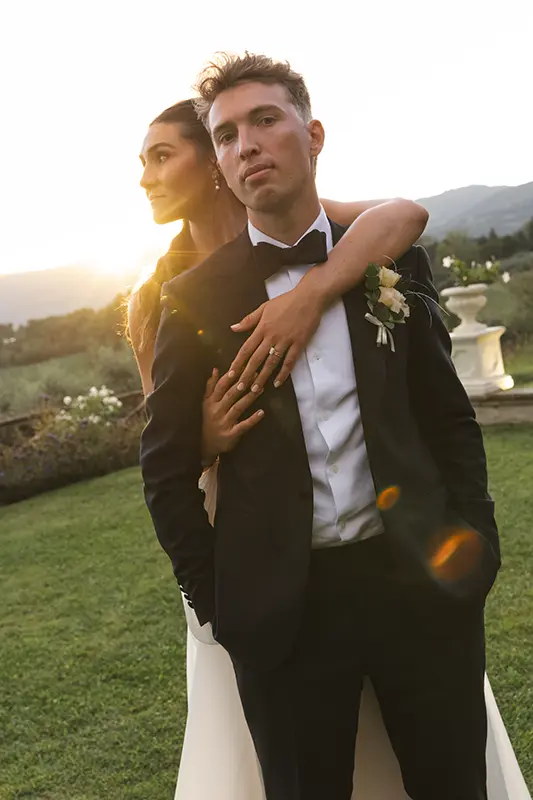 Bride hugs groom from behind at sunset
