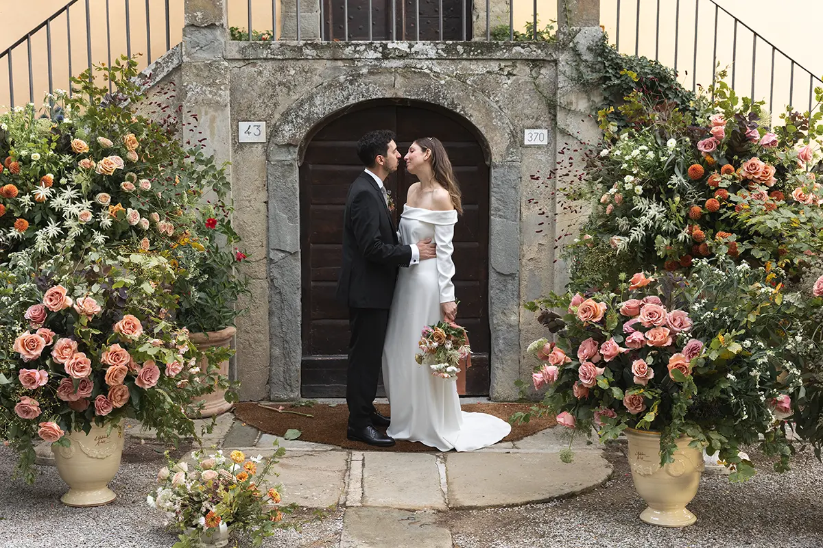 A newlywed couple embraces in front of the gate of a villa in Tuscany, surrounded by flowers.