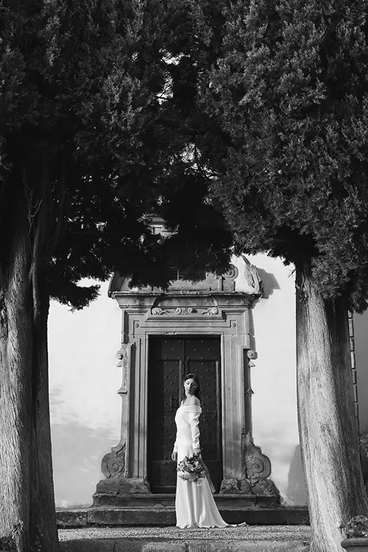 Portrait of the bride in front of the door of the old church of the Relais il Falconiere framed by cypress trees